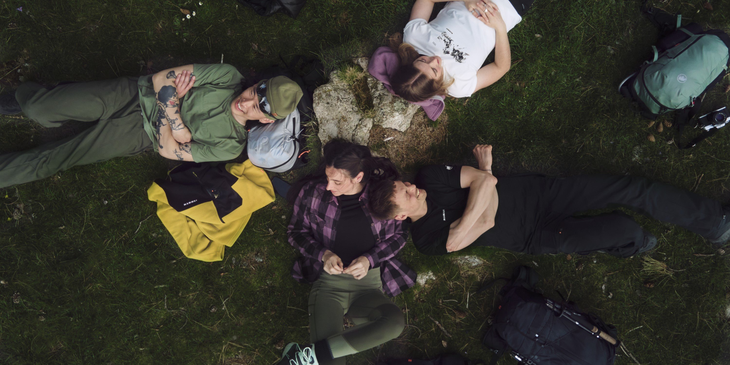 Four adventurers wearing Mammut gear lying on grass in a circle, heads together, with backpacks and hiking equipment scattered nearby.