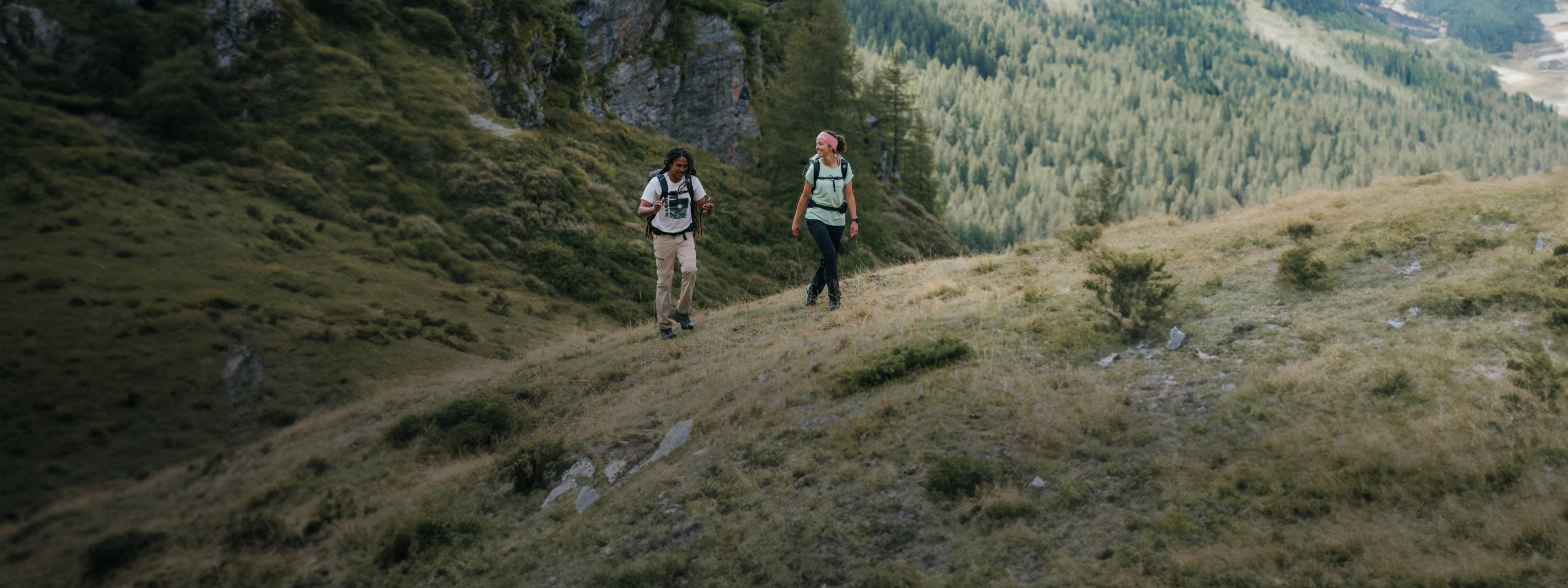 Two people wearing Mammut gear hiking on a scenic mountain trail surrounded by lush trees, with a picturesque valley in the background.