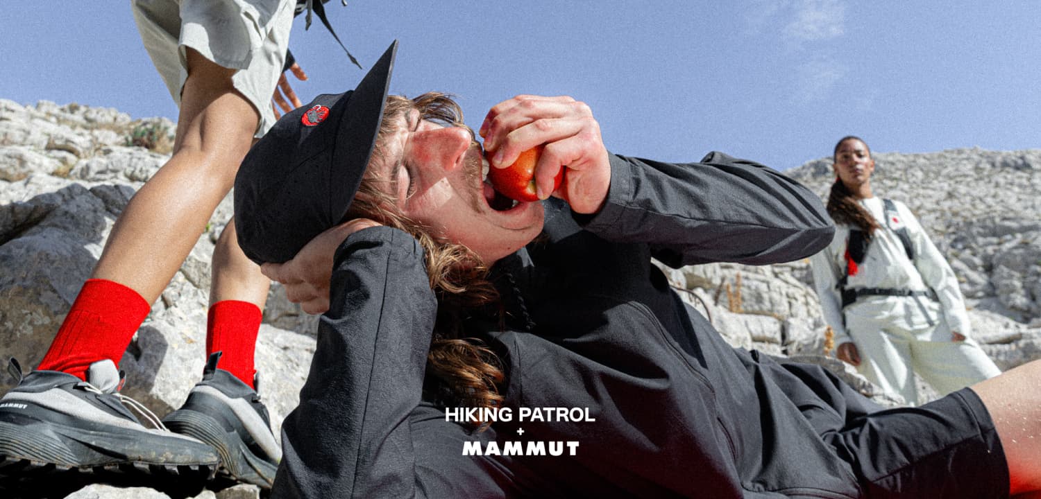 Hiker in Mammut gear lies on rocky ground, eating an apple as others stand nearby, with mountains in the background.